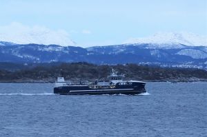 Blå brønnbåt seilende midt i bildet. I bakgrunnen er det fjell. Grått vær, stille sjø